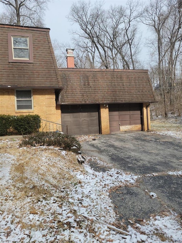 view of snowy exterior with driveway, roof with shingles, an attached garage, a chimney, and brick siding