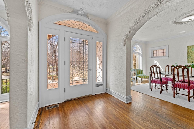 entryway with visible vents, hardwood / wood-style floors, arched walkways, crown molding, and a textured wall