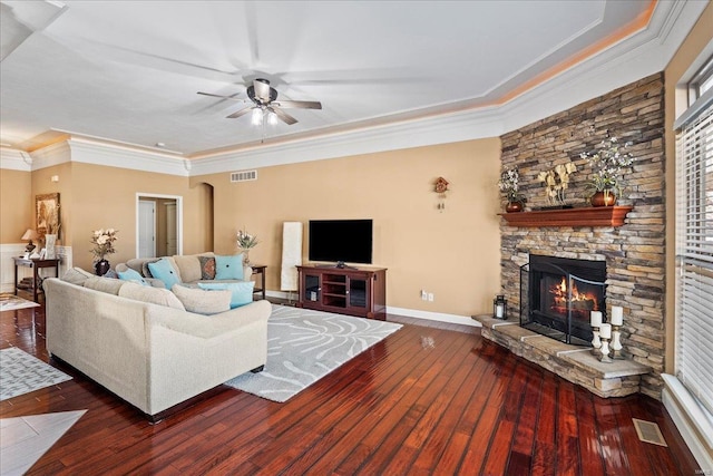 living area featuring visible vents, crown molding, dark wood-type flooring, a stone fireplace, and a ceiling fan