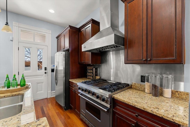 kitchen with light stone counters, stainless steel appliances, hanging light fixtures, wall chimney exhaust hood, and light wood-type flooring