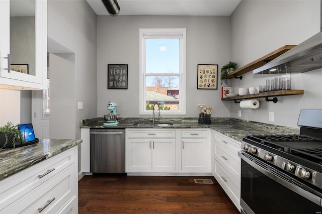 kitchen with dark stone countertops, white cabinets, stainless steel appliances, wall chimney exhaust hood, and a sink