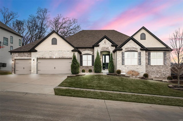 french country home featuring a lawn, driveway, an attached garage, a shingled roof, and brick siding