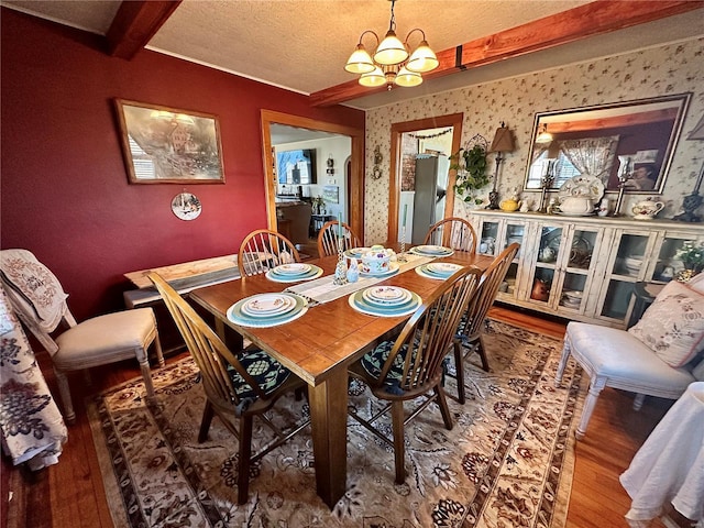 dining space featuring wallpapered walls, beamed ceiling, wood finished floors, a notable chandelier, and a textured ceiling