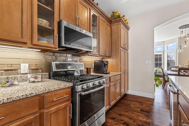 kitchen featuring brown cabinets, dark wood-type flooring, light stone counters, stainless steel appliances, and decorative backsplash