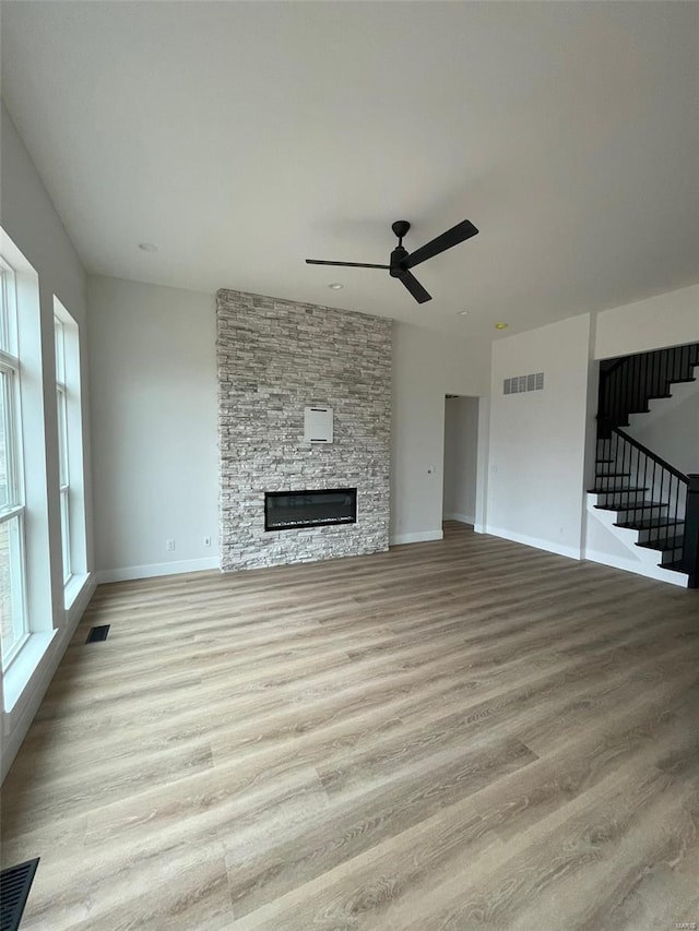 unfurnished living room featuring a stone fireplace, visible vents, a ceiling fan, light wood-style flooring, and stairs