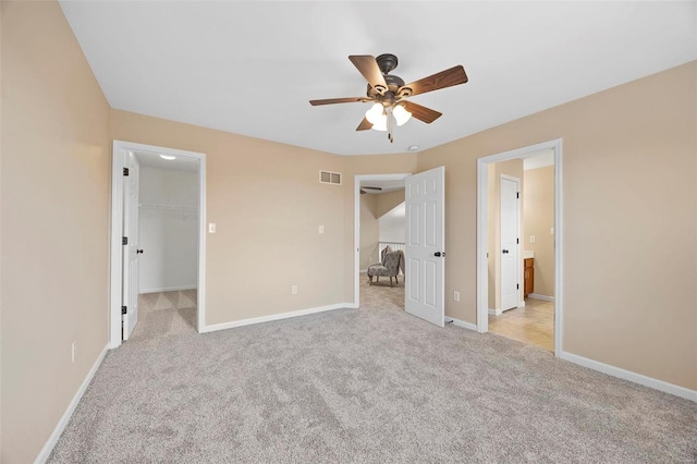 unfurnished bedroom featuring a walk in closet, visible vents, baseboards, light colored carpet, and ceiling fan