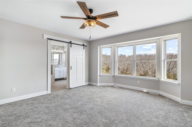 unfurnished bedroom featuring connected bathroom, baseboards, light colored carpet, a barn door, and a ceiling fan