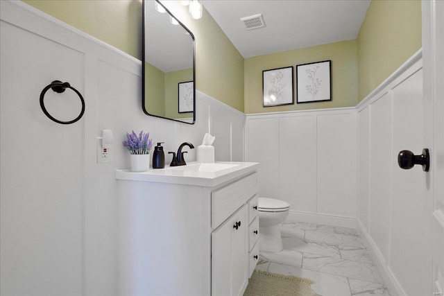 bathroom featuring visible vents, toilet, a decorative wall, marble finish floor, and vanity