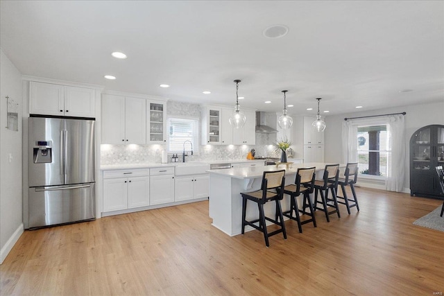 kitchen featuring a kitchen island with sink, a sink, light countertops, stainless steel refrigerator with ice dispenser, and wall chimney range hood