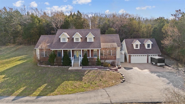 cape cod-style house with a forest view, a front lawn, concrete driveway, covered porch, and an outdoor structure