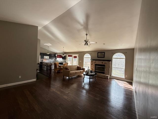 living room featuring a ceiling fan, wood finished floors, baseboards, vaulted ceiling, and a brick fireplace