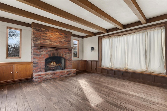 unfurnished living room featuring beamed ceiling, a brick fireplace, hardwood / wood-style floors, and wainscoting