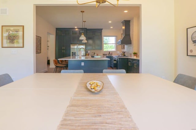 kitchen featuring an island with sink, stainless steel refrigerator with ice dispenser, wall chimney range hood, a kitchen breakfast bar, and light countertops