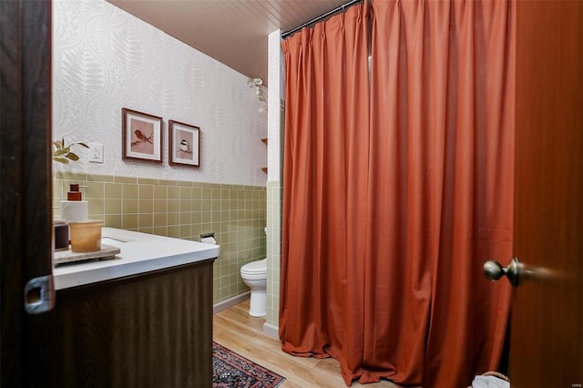 bathroom featuring wood finished floors, a wainscoted wall, wallpapered walls, tile walls, and toilet