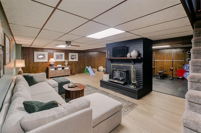 living room featuring a drop ceiling, wood finished floors, wooden walls, ceiling fan, and a wood stove