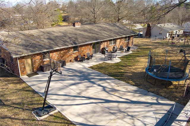 exterior space with brick siding, a trampoline, roof with shingles, a yard, and a patio area