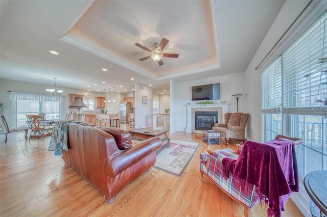 living area featuring light wood finished floors, a tray ceiling, recessed lighting, a high end fireplace, and ceiling fan with notable chandelier