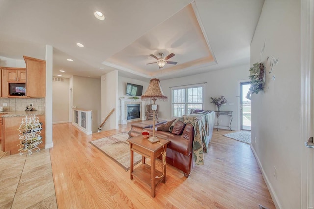 living room featuring visible vents, a glass covered fireplace, light wood finished floors, a raised ceiling, and ceiling fan