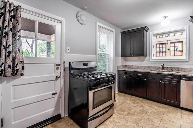 kitchen with light tile patterned floors, light stone countertops, a sink, appliances with stainless steel finishes, and wainscoting