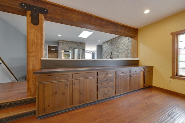 kitchen featuring brown cabinetry, wood finished floors, baseboards, beam ceiling, and stainless steel appliances