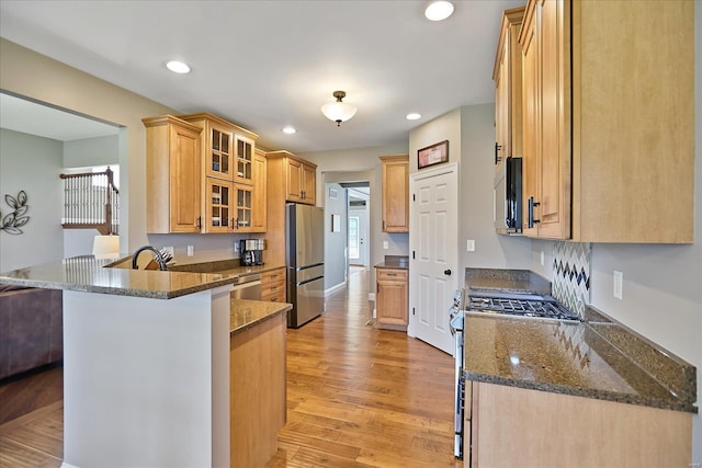 kitchen featuring light wood-type flooring, dark stone counters, appliances with stainless steel finishes, a peninsula, and glass insert cabinets