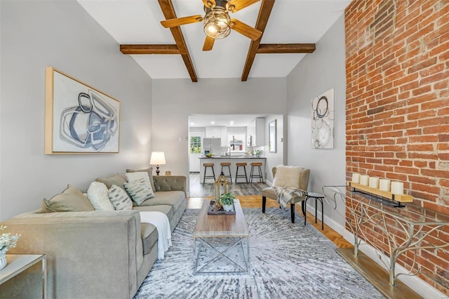 living room featuring brick wall, baseboards, beam ceiling, wood finished floors, and a ceiling fan