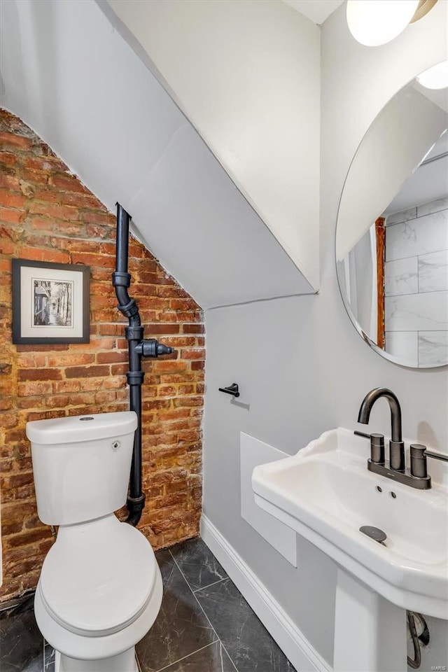 half bathroom featuring brick wall, lofted ceiling, a sink, toilet, and marble finish floor