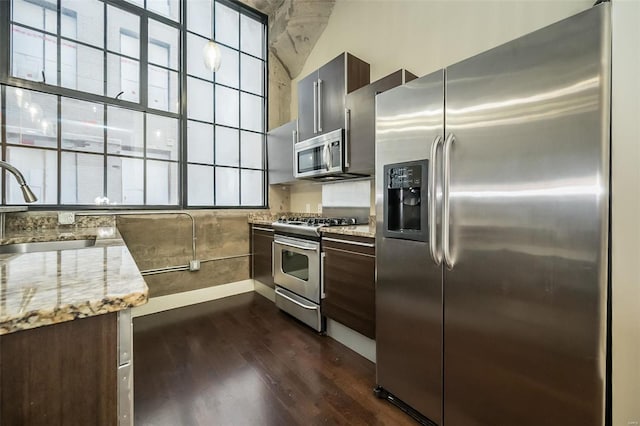 kitchen with a sink, light stone counters, dark brown cabinetry, appliances with stainless steel finishes, and dark wood-style flooring