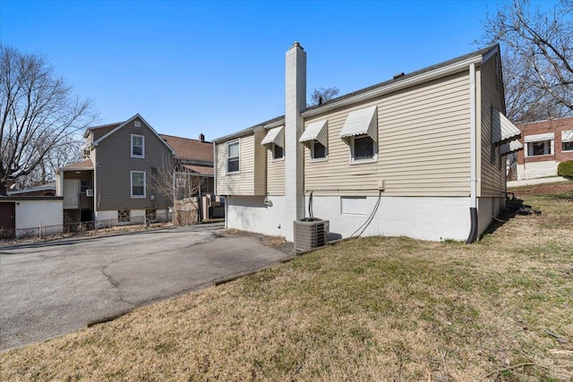 rear view of house with aphalt driveway, central AC unit, a chimney, and a lawn