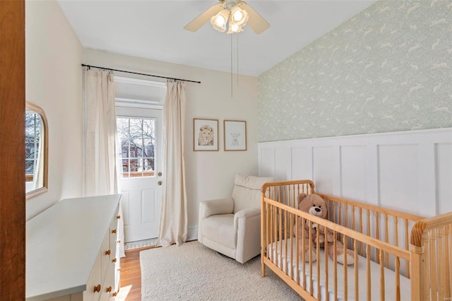 bedroom with wood finished floors, a wainscoted wall, wallpapered walls, ceiling fan, and a nursery area