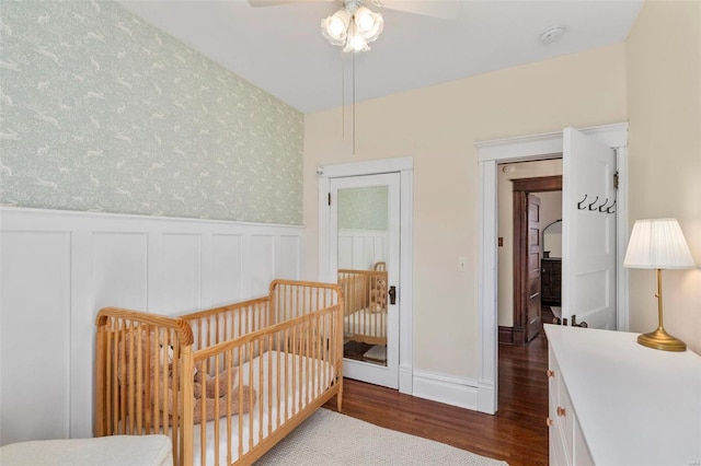 bedroom with dark wood finished floors, a nursery area, a wainscoted wall, and wallpapered walls