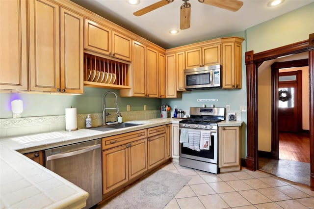 kitchen featuring tile countertops, light tile patterned floors, a ceiling fan, a sink, and appliances with stainless steel finishes