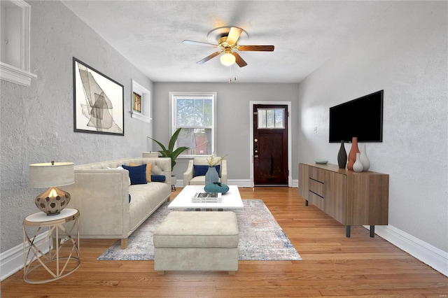 living area featuring a textured wall, baseboards, ceiling fan, and light wood finished floors