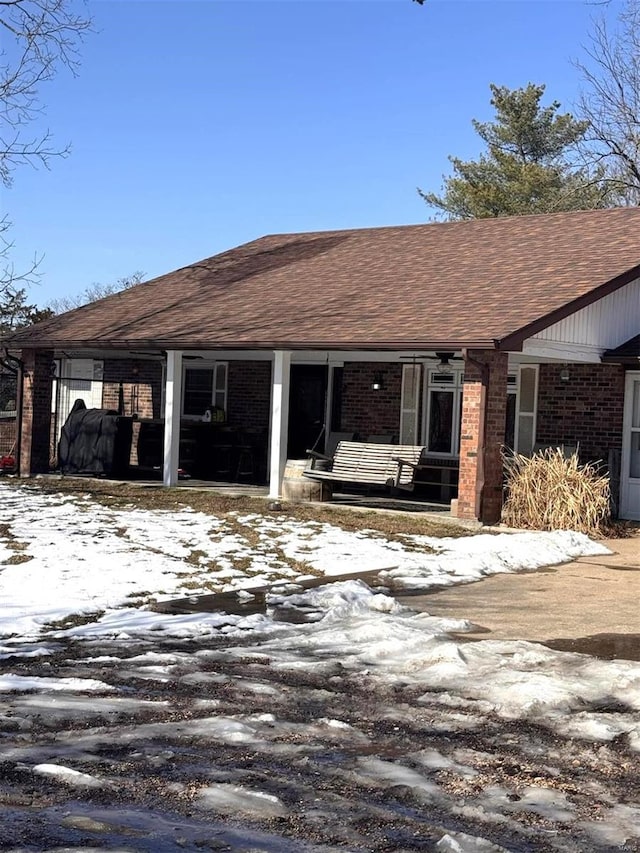 snow covered house with brick siding, covered porch, and roof with shingles