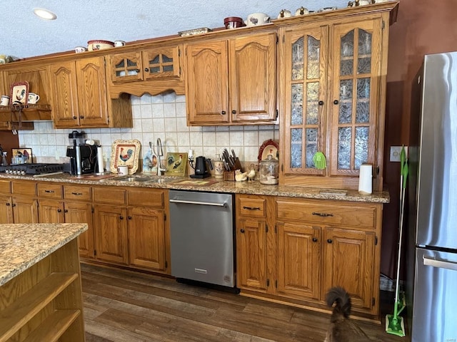 kitchen with glass insert cabinets, decorative backsplash, brown cabinetry, stainless steel appliances, and dark wood-style flooring