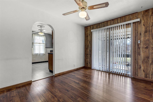 empty room with baseboards, ceiling fan, wood walls, hardwood / wood-style flooring, and arched walkways