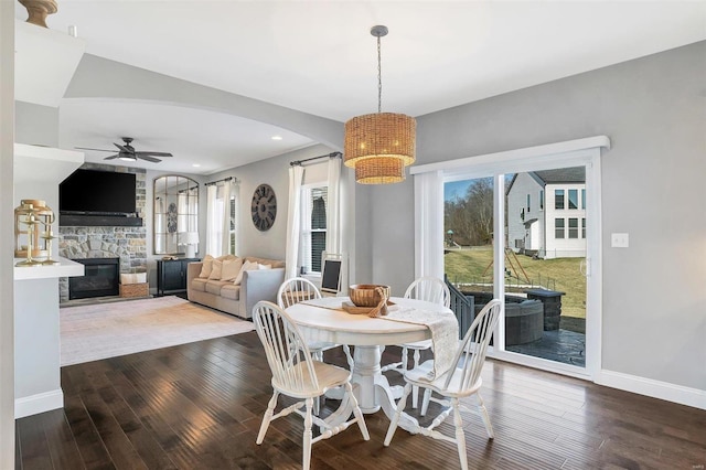 dining space with dark wood-style floors, baseboards, arched walkways, ceiling fan, and a stone fireplace