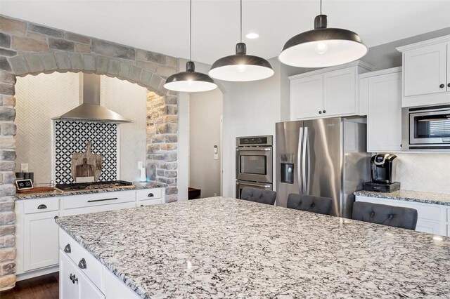 kitchen featuring tasteful backsplash, stainless steel appliances, wall chimney exhaust hood, white cabinets, and light stone countertops