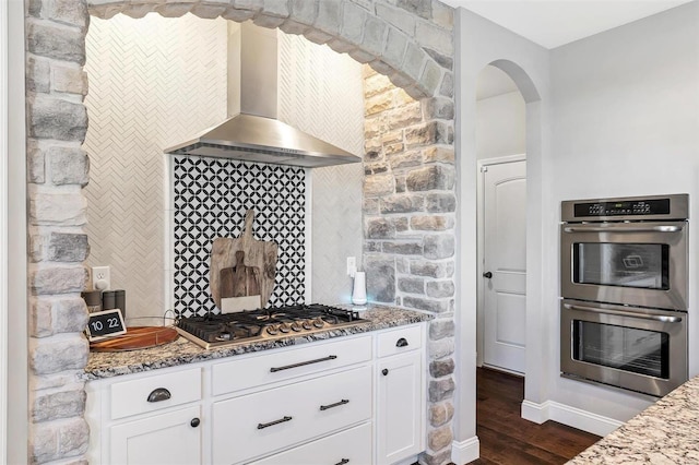 kitchen featuring wall chimney range hood, light stone countertops, appliances with stainless steel finishes, dark wood-style floors, and white cabinetry