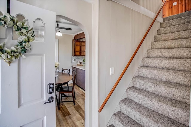 stairway featuring wood finished floors, arched walkways, ornamental molding, ceiling fan, and wood walls