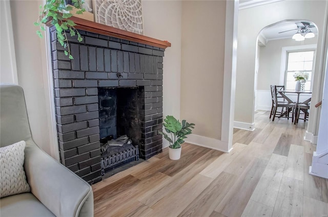 living room featuring a brick fireplace, ceiling fan, ornamental molding, wood finished floors, and arched walkways