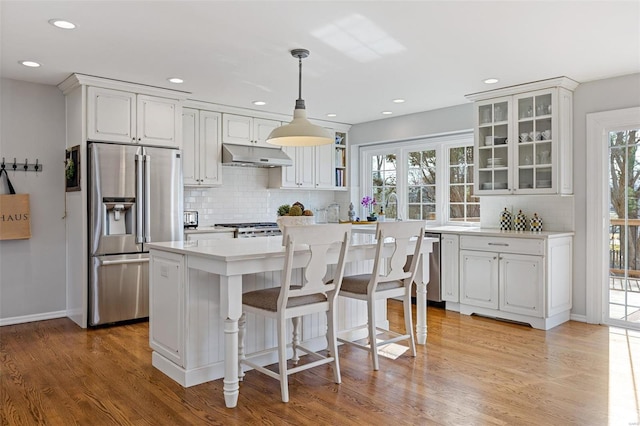 kitchen featuring under cabinet range hood, stainless steel appliances, light wood-style floors, light countertops, and glass insert cabinets