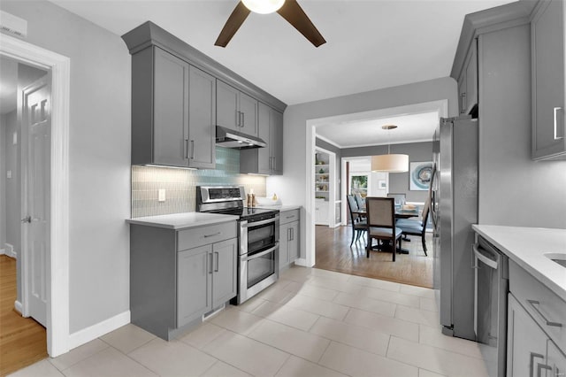 kitchen with gray cabinetry, decorative backsplash, light countertops, under cabinet range hood, and appliances with stainless steel finishes