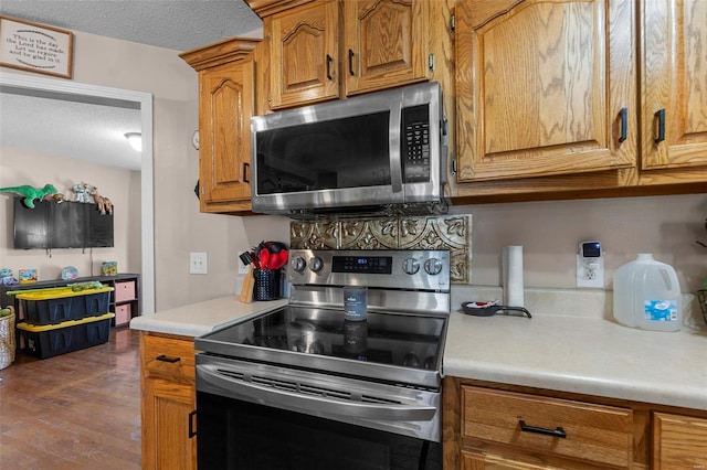 kitchen with a textured ceiling, dark wood-style floors, stainless steel appliances, brown cabinetry, and light countertops