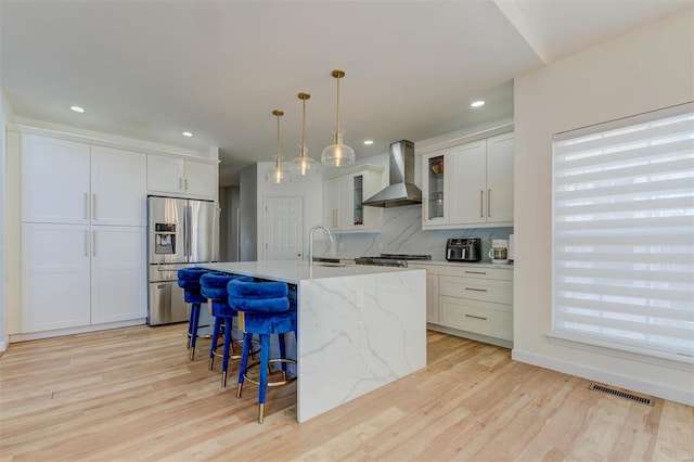 kitchen featuring backsplash, wall chimney range hood, a kitchen bar, light wood-style floors, and stainless steel refrigerator with ice dispenser