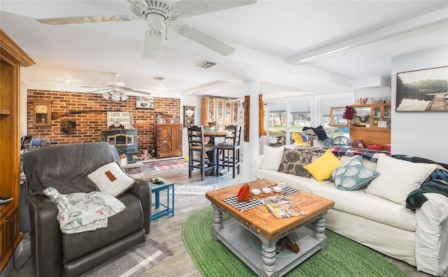 living area with a wood stove, beam ceiling, brick wall, visible vents, and a ceiling fan