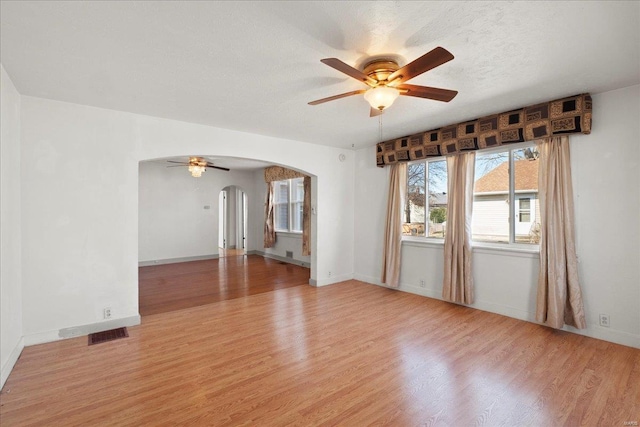 empty room featuring a textured ceiling, arched walkways, light wood finished floors, baseboards, and ceiling fan