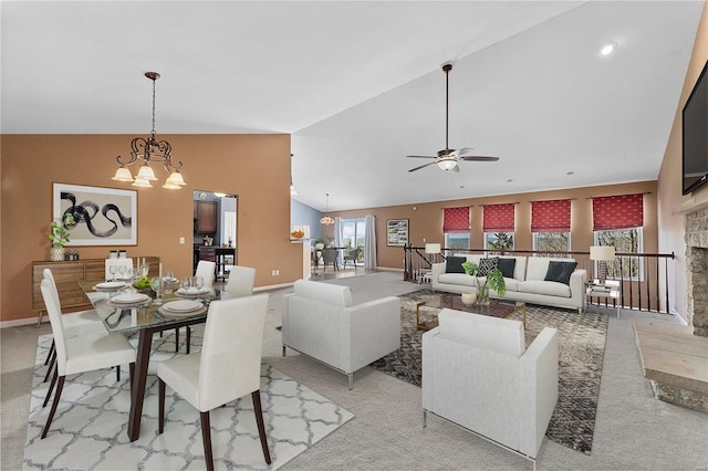 dining room with baseboards, high vaulted ceiling, a stone fireplace, ceiling fan with notable chandelier, and light colored carpet