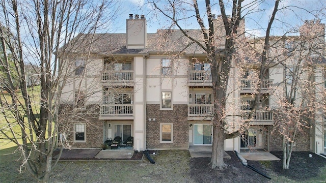 rear view of house with a patio, a shingled roof, stucco siding, a chimney, and brick siding