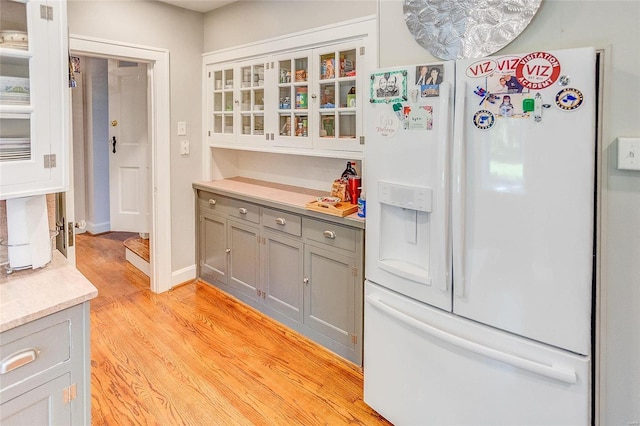 kitchen featuring light wood finished floors, gray cabinetry, white fridge with ice dispenser, light countertops, and glass insert cabinets